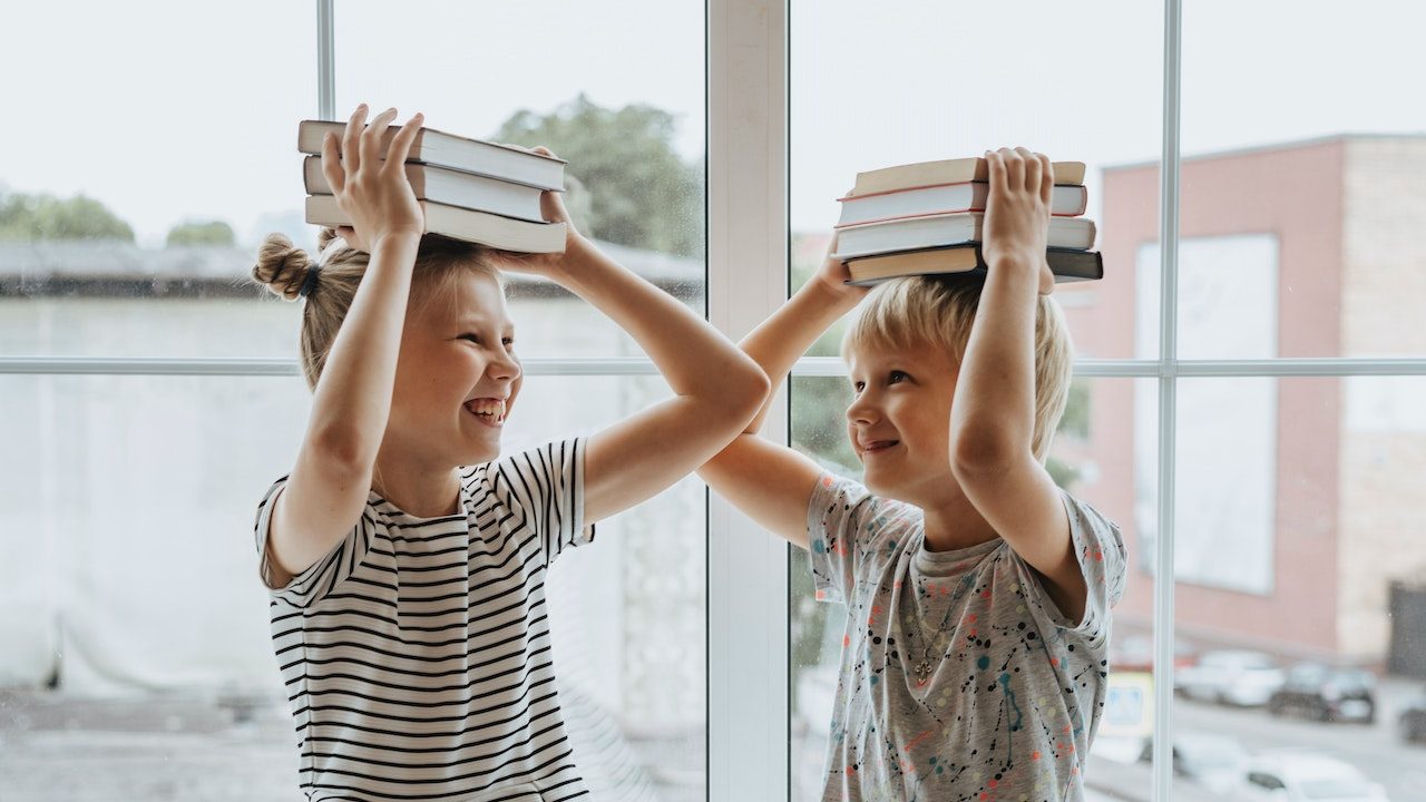 Zwei Kinder tragen mehrere Bücher auf ihren KKöpfen und lachen dabei.